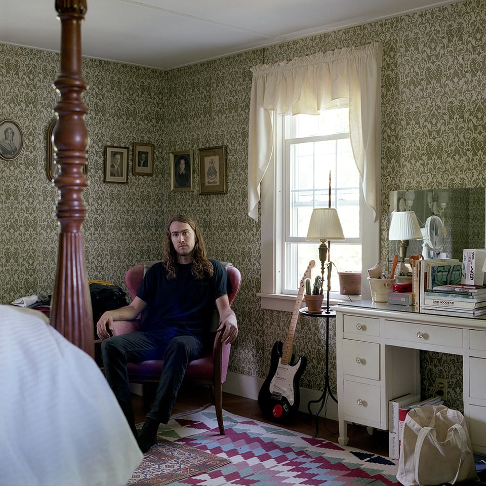 Ein Mann mit langen Haaren in einem Schlafzimmer mit grüner, gemusterter Tapete.