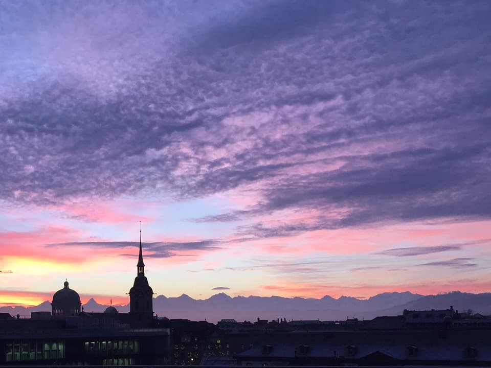 Unten im Bild die Silhouette der Stadt Bern inklusive dem Bundeshaus. Dahinter die Riesen des Berner Oberlands. Darüber ein malerisches Bild aus Wolkenfeldern in rosa, orange und violetten Farbtönen.