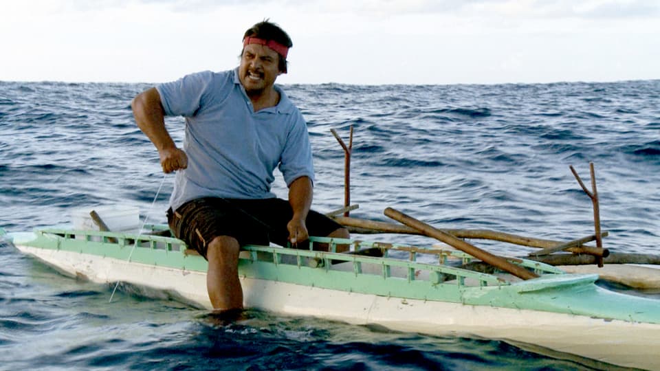 Ein Mann sitzt in Shorts und T-Shirt auf einem Kanu und zieht an einer gespannten Fischerleine.