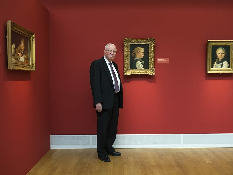 Blocher steht vor einer roten Wand mit Gemälden