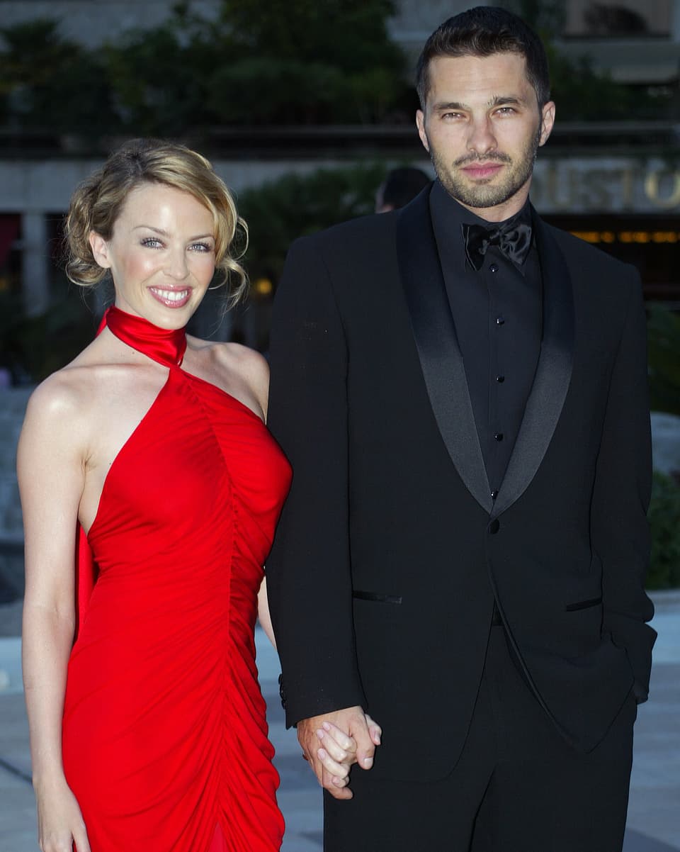 Kylie Minogue im roten Kleid hält Händchen mit einem Mann im schwarzen Anzug. 