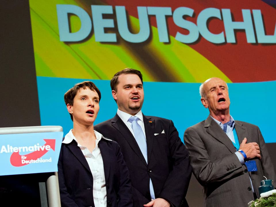 Zwei Politikerinnen und zwei Politiker nebeneinander stehend, singend, hinter ihnen ist eine deutsche Flagge projiziert, darauf steht gross "Deutschland".