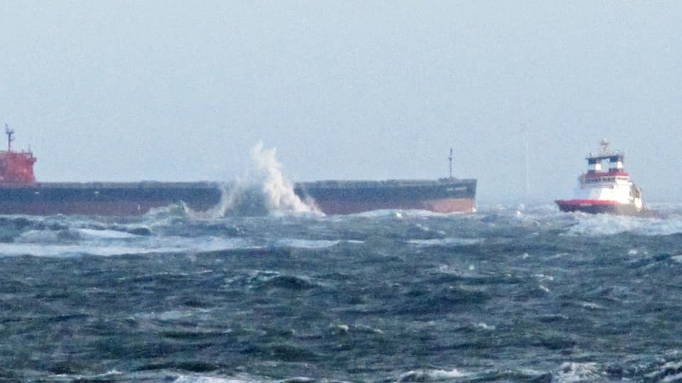 Der auf Grund gelaufene Frachter, an dem die Wellen hochgehen und der Schlepper.