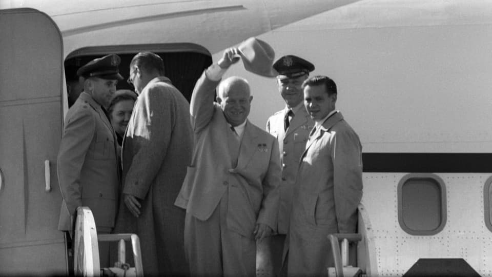 In der Mitte des Bildes winkt Nikita Chruschtschow mit seinem Hut, nachdem er aus einem Flugzeug ausgestiegen ist. 