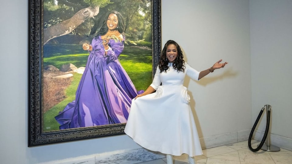 Eine afroamerikanische Frau in einem weissen Kleid steht vor einem Porträt von ihr in einem lilafarbenen Kleid.