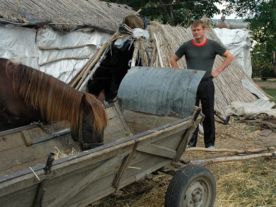 Ein Mann steht bei einem Holzwagen, aus dem ein Pferd Heu frisst.