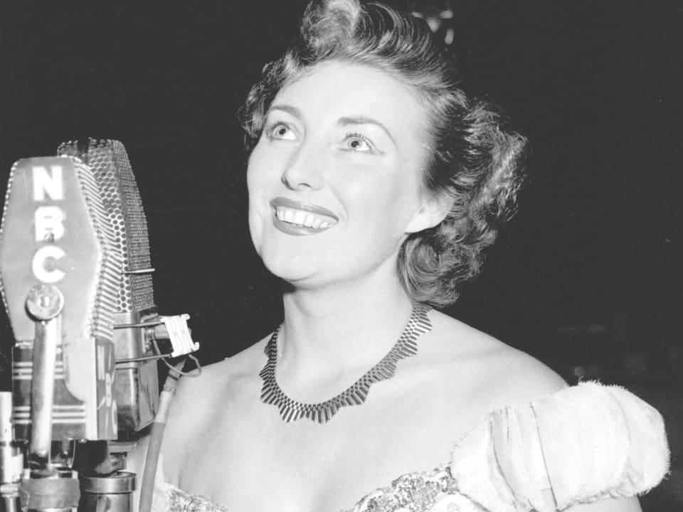 Ein Schwarzweiss-Bild einer Frau, die vor einem Mikorfon steht.
