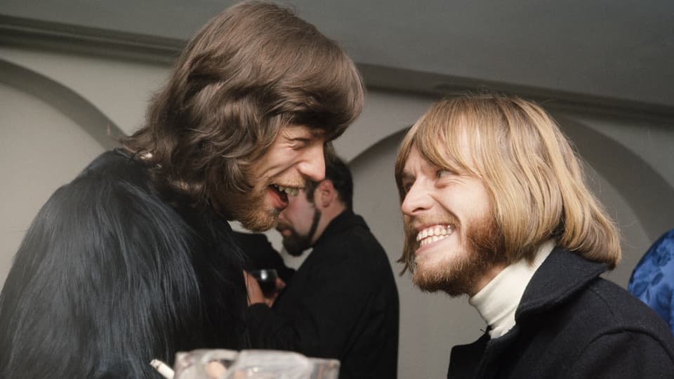 Zwei Männer mit längeren Haaren und Bärten. Jagger trägt einen Mantel mit langem Fell.