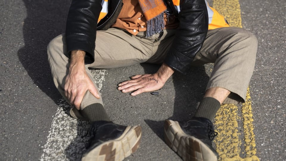 Mann sitzt auf einer Strasse. Eine Hand ist am Boden festgeklebt.