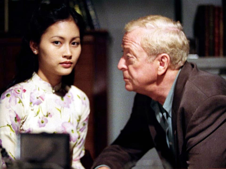 Filmszene, junge asiatische Frau, die in die Kamera blickt, daneben ein älterer Mann, der sie anschaut.