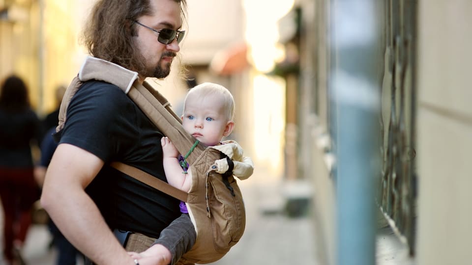 Ein Mann mit langen Haaren und Sonnenbrille trägt einen kleinen Jungen im Tragtuch auf dem Bauch.