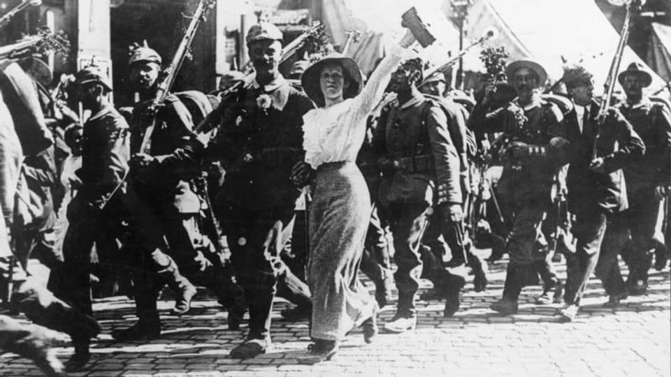 Eine Gruppe Soldaten. Eine Frau hängt sich beim einen Soldat ein und hebt ihren linken Arm triumphierend in die Höhe.