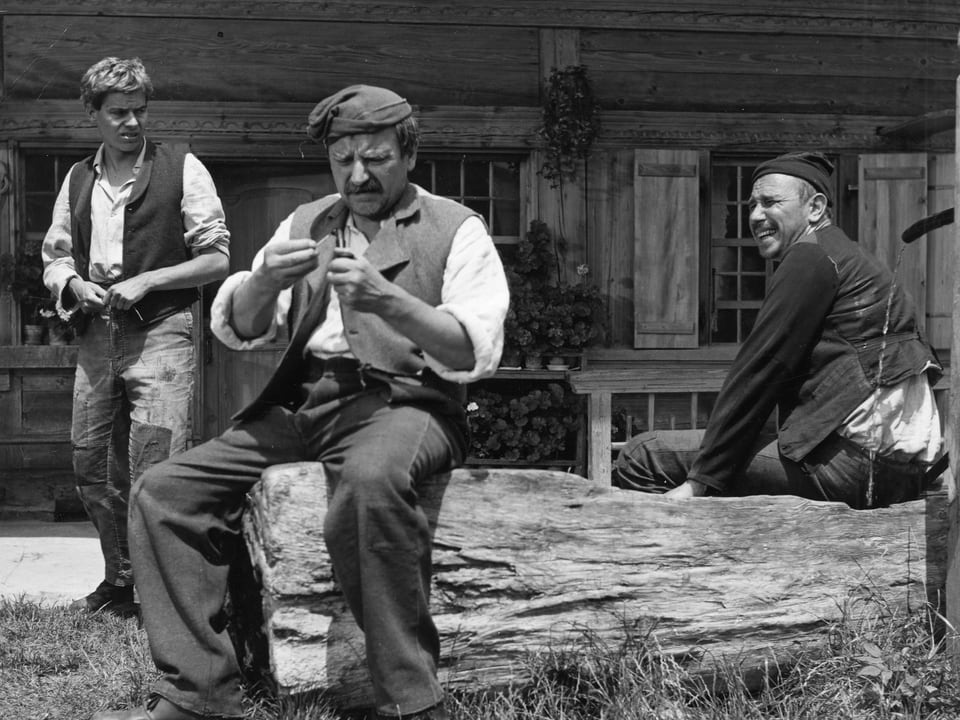 Zwei Männer sitzen auf einem Baumstamm, der auch als Brunnentrog dient. Der Mann links betrachtet konzentriert einen Gegenstand in seinen Händen. Links hinter ihm steht ein dritter, etwas jüngerer Mann.