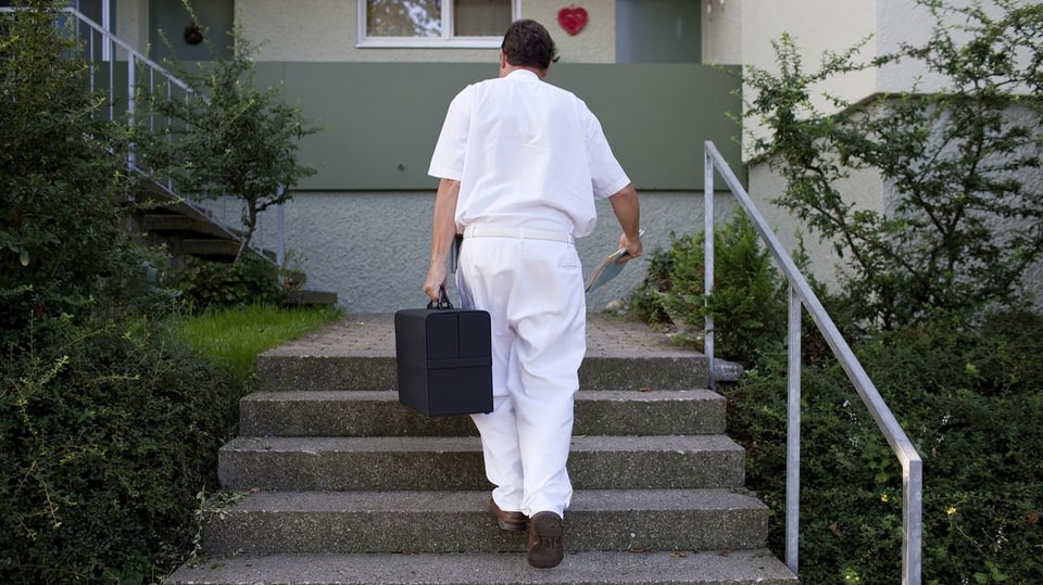 Ein Mann in weissem Arztgewand und mit scharzem Arztkoffer steigt Treppen zu einem Wohnhaus hoch