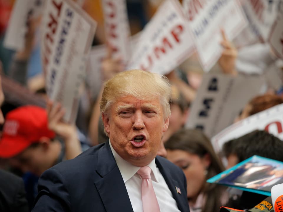 Donald Trump mit Schnute vor Anhängern mit Wahlplakaten.