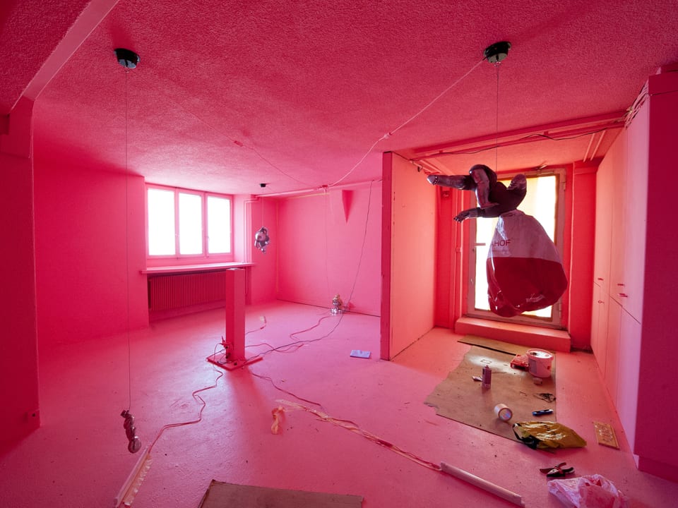 Ein mit Pink ausgeleuchteter Raum, in dem Puppen und nicht klar definierbare Objekte an Schnüren aufgehängt sind und am Boden liegen.