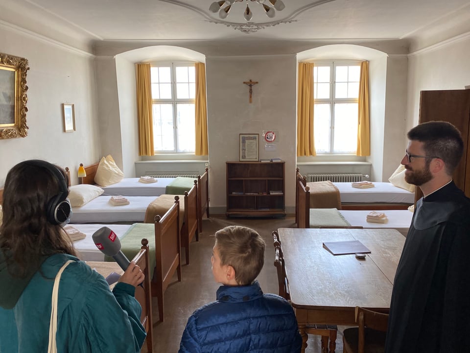 Dania, Nick und Pater Philipp schauen in ein Zimmer, in dem an beiden Wänden je vier Betten stehen.