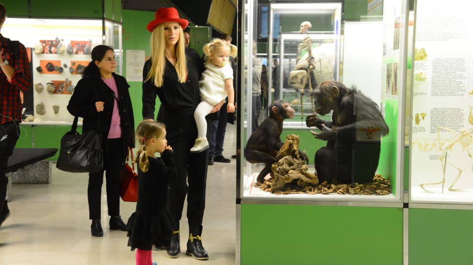 Eine Frau steht mit ihren zwei Kindern vor einer Glasbox, in der zwei ausgestopfte Affen sitzen. 