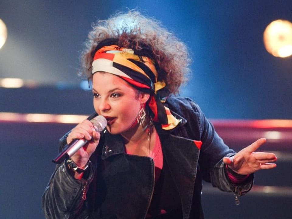 Woman with curly hair singing on stage.  She is wearing a colorful hairband.