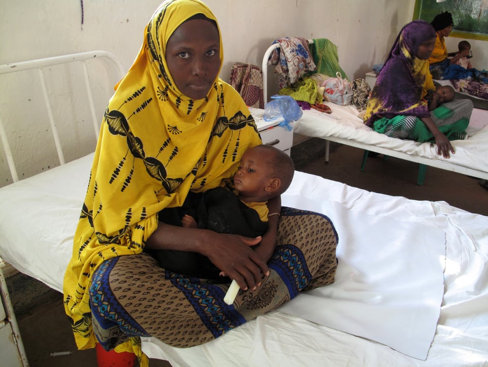 Frau in gelbem Gewand auf einem Spitalbett. Sie hält ein abgemagertes Kind in den Armen.