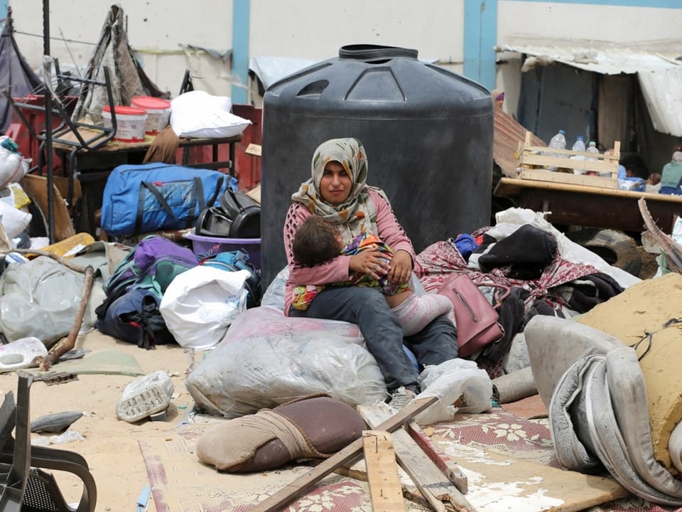 Frau mit Kind sitzt zwischen Habseligkeiten in einem behelfsmässigen Lager.