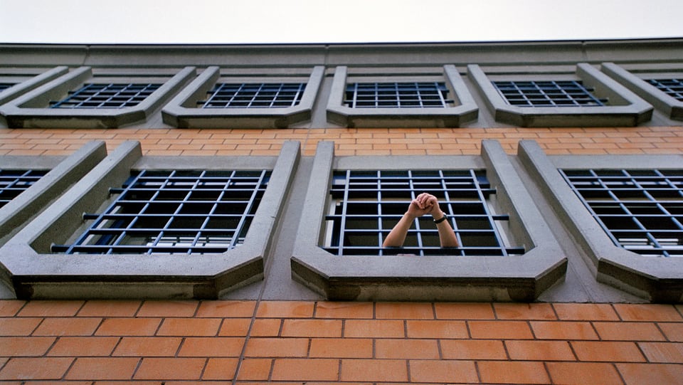 Gefängnis von aussen, aus einem der vergitterten Fenstern hält eine Person ihre Arme heraus.