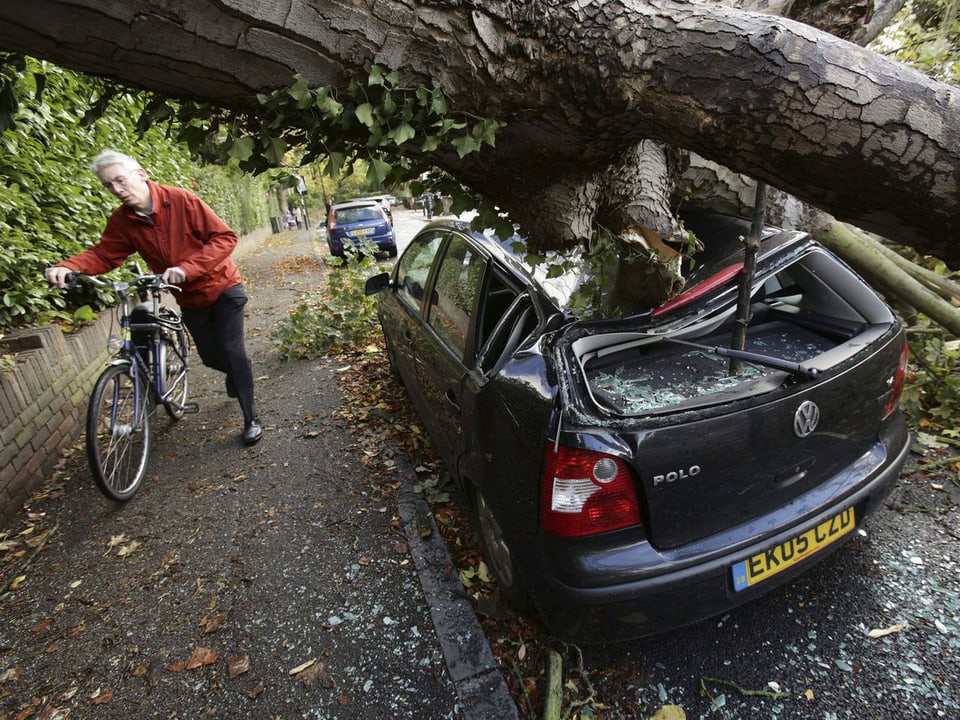 Ein ausgerissener Baum liegt auf einem Auto. Auf den Trottoir schiebt ein Mann sein Velo vorbei. (keystone)