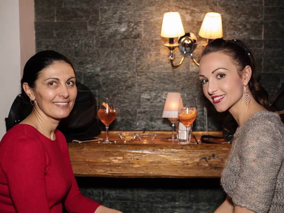 Zwei Frauen sitzen an einer Bar vor zwei Weingläsern. Sie schaun von der Seite lächelnd in die Kamera. Beide haben lange, dunkle Haare, die zu einem Pferdeschwanz zurückgebunden sind. Die rechts trägt einen roten Pulli, die andere einen beigen Strickpullover.
