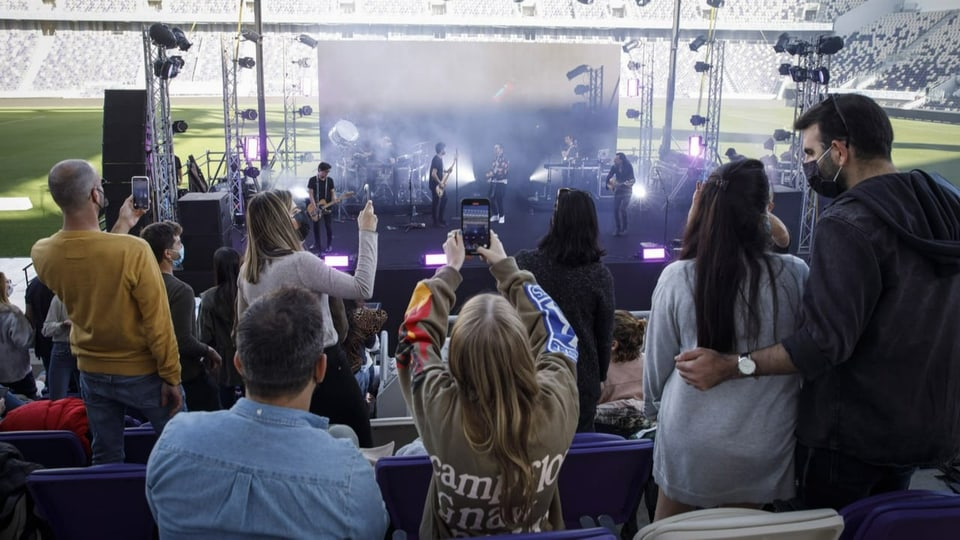 Im Hintergrund eine Band auf einer Bühne in eine Stadion. Davor Zuschauerinnen, die mit den Handy das Konzert filmen.