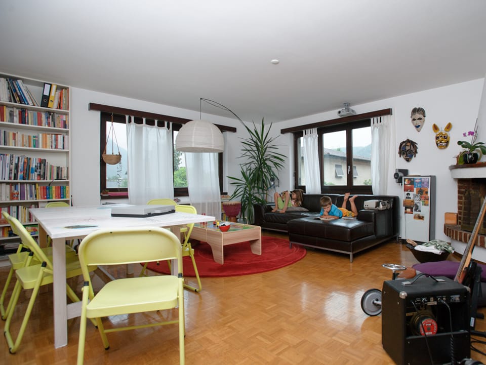 Blick ins grosse Wohnzimmer mit braunem Ecksofa aus Leder, um einen weissen Tisch stehen zartgelbe Stühle.