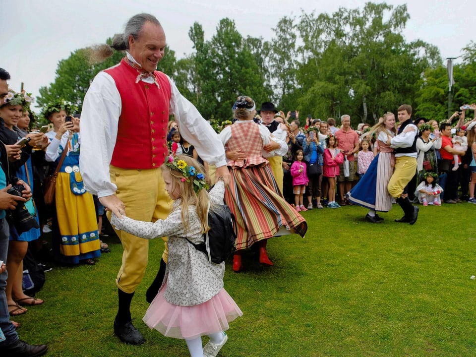 Ein Mann in einer Tragt tanzt  mit einem Mädchen auf einer Wiese. Viele Leute schauen zu.