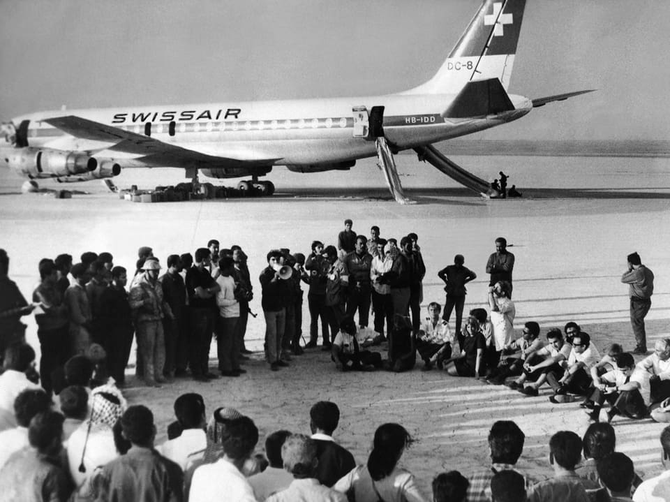 Schwarz-Weiss Aufnahme einer Ansammlung von Menschen, die im Kreis stehen, im Hintergrund ein Flugzeug «Swissair».