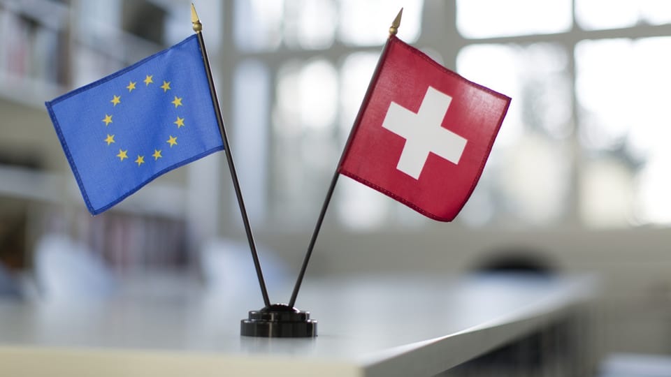 Links blau-gelbe EU-Flagge, rechts rot-weisse Schweizflagge, stehen auf weissem Tisch.