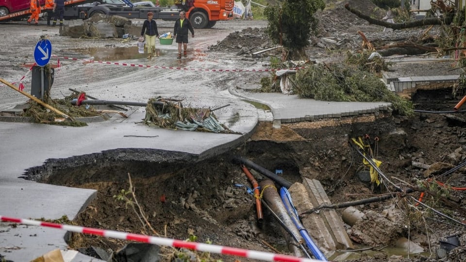 Menschen tragen in Schuld ihr Hab und Gut weg. Im Vordergrund eine eingebrochene und weggespülte Strasse.