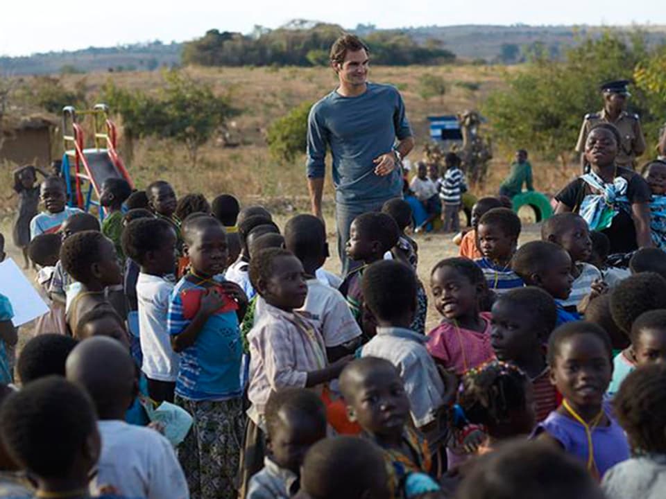 Roger Federer ist in Malawi. Er steht auf einem Soielplatz mit vielen malawischen Kindern. Im Hintergrund sieht man eine farbige, kleine Rutsche.
