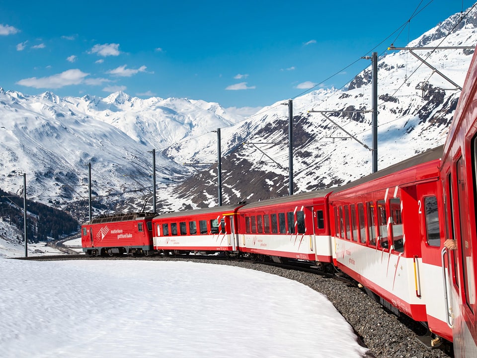 Ein rot-weisser Zug fährt durch eine schneebedeckte Landschaft. Im Hintergrund sieht man einen Teil der Alpen und strahlend blauen Himmel.