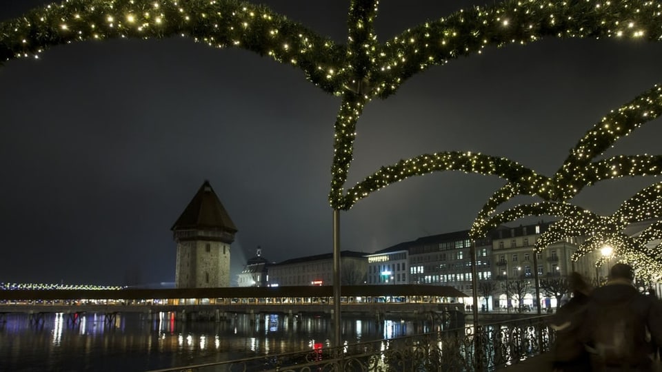 Menschen gehen auf einer Brücke mit grünen Kränzen und Lichterketten. Im Hintergrund ein runder Turm mit Holzdach.