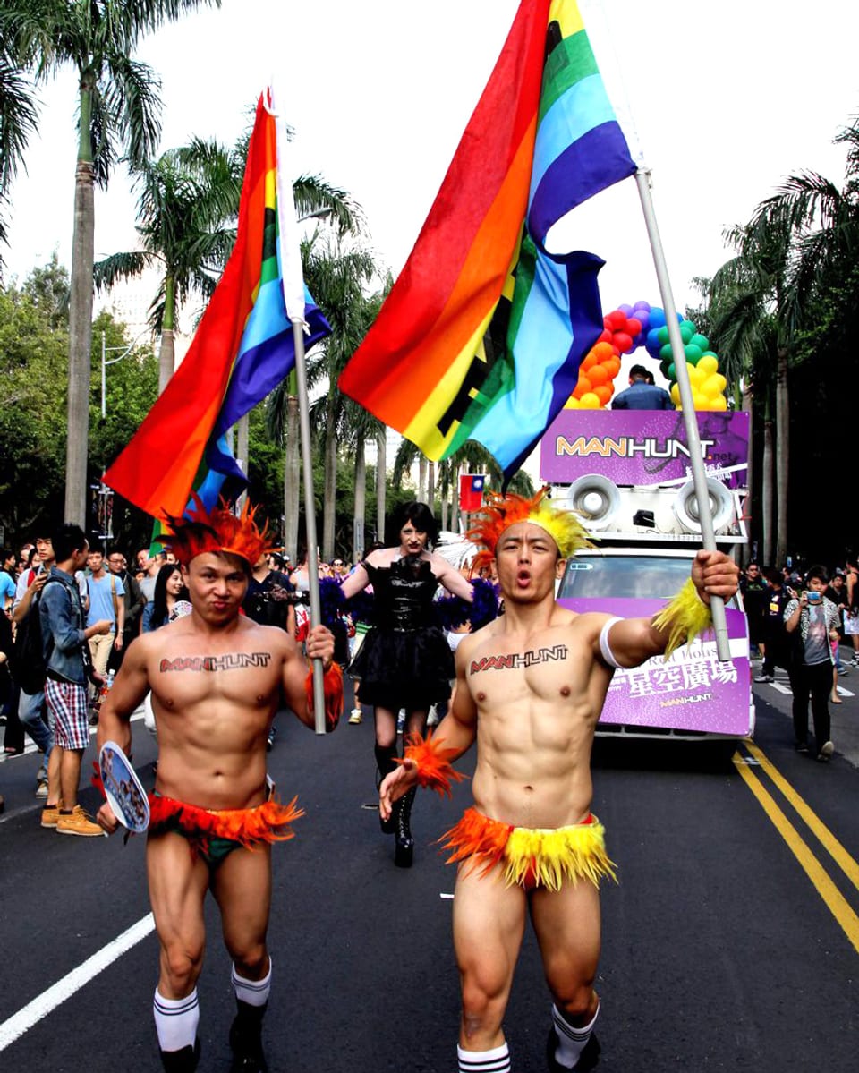 Zwei Männer in bunten Unterhosen halten je eine regenbogenfarbene Flagge in die Luft, dahinter eine Parade mit vielen Menschen.
