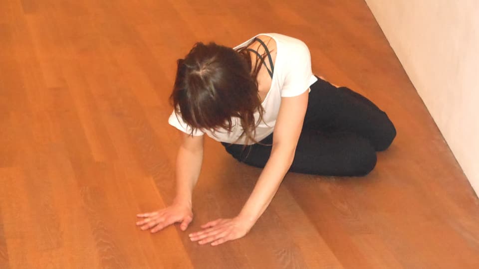 Eine Frau in Gymnastikhose und weissem T-Shirt sitzt leicht verdreht auf Holzboden.