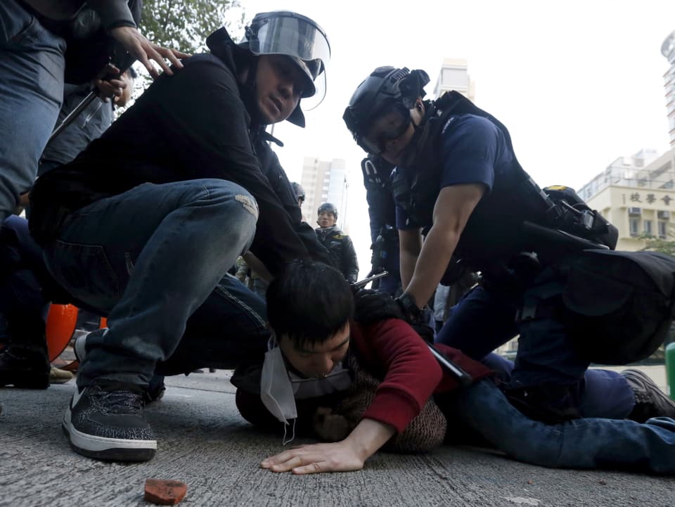 Zwei Polizisten in voller Montur drücken einen jungen Mann zu Boden.
