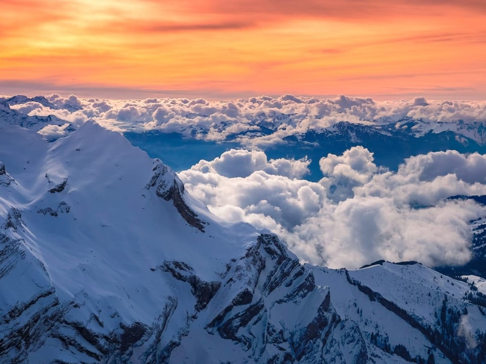 Gelb bis rote Schleierwolken über verschneiten Alpengipfeln mit Wolken in den Tälern. 