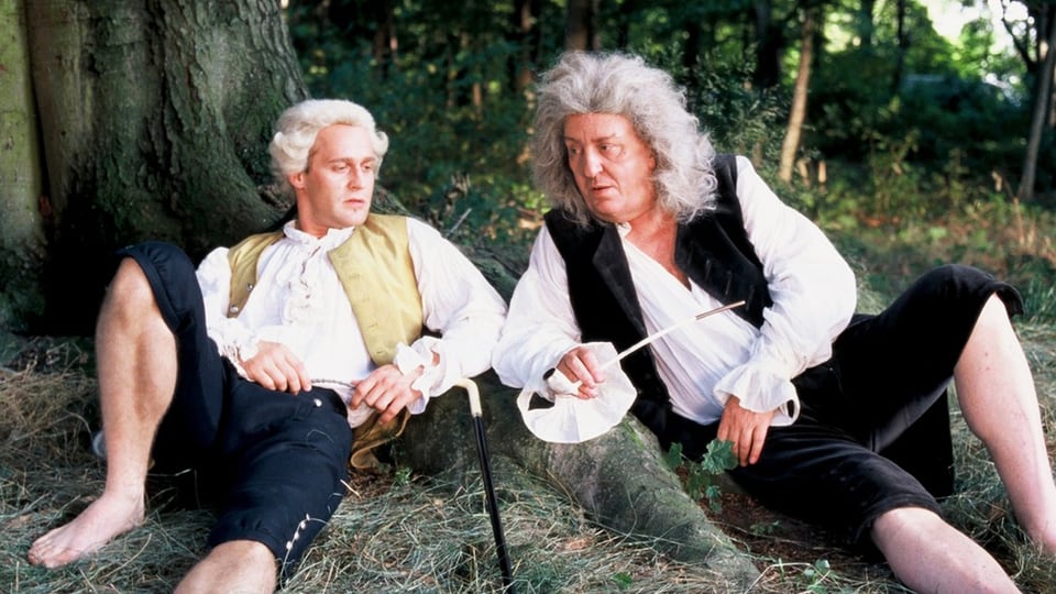 Zwei Männer in klassischen Kleidern lehnen an einer Baumwurzel.