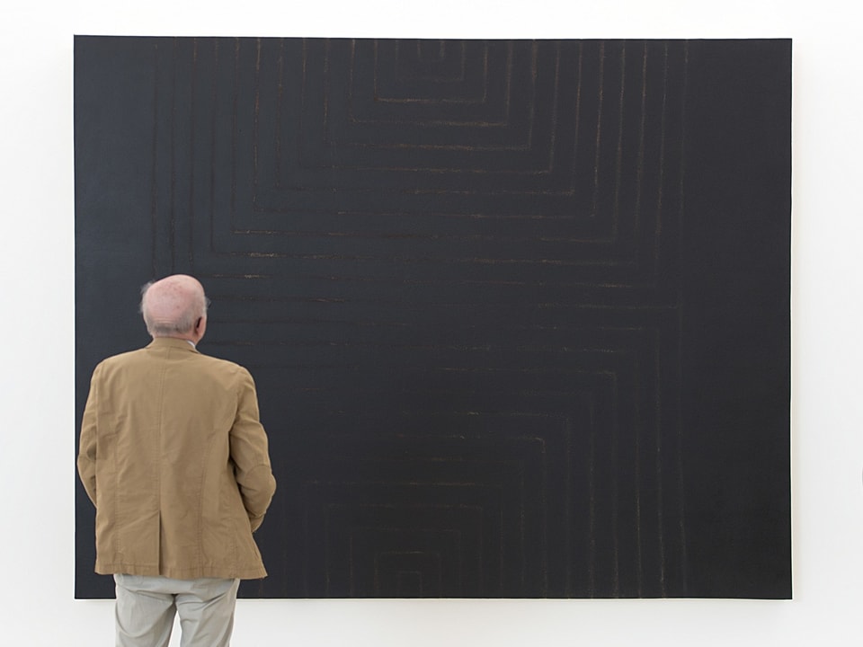 Ein alter Mann mit hellbraunem Jackett steht vor einem grossen, schwarzen Bild.