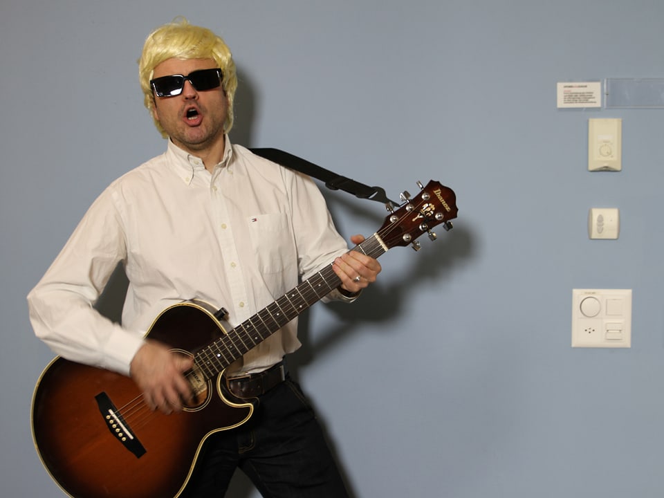 Der Moderator trägt eine blonde Perücke, eine dunkle Sonnenbrille und spielt Gitarre.