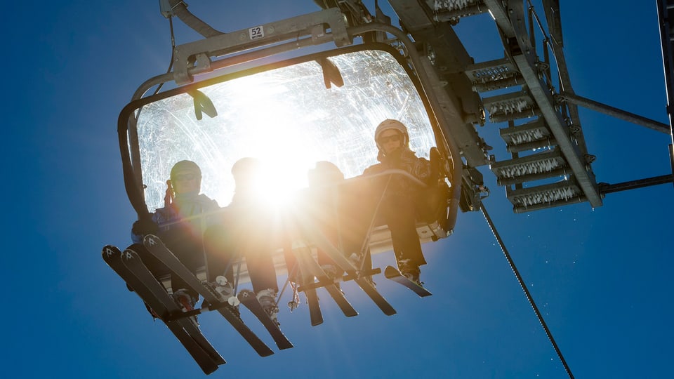 Vier Skifahrer in einer Gondel einer Sesselbahn, durch deren Haube die Sonne scheintl