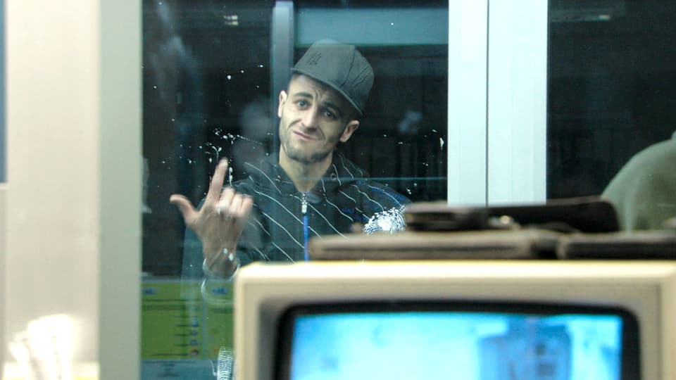 Szene aus dem Film «La Forteresse» mit einem Mann hinter Glas, der einen Hut trägt.