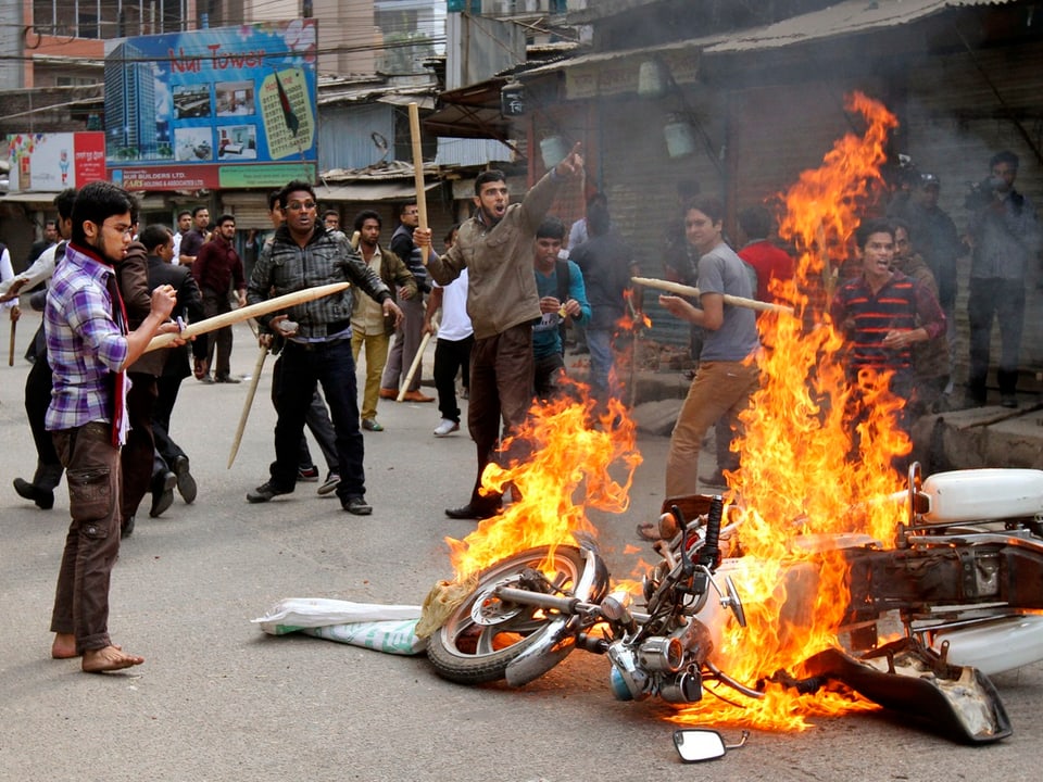 Wütende Männer stehen um ein Motorrad, das in Brand gesteckt wurde.