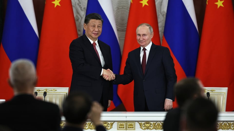 Xi und Putin geben sich die Hand, sie stehen vor Flaggen Chinas und Russlands.