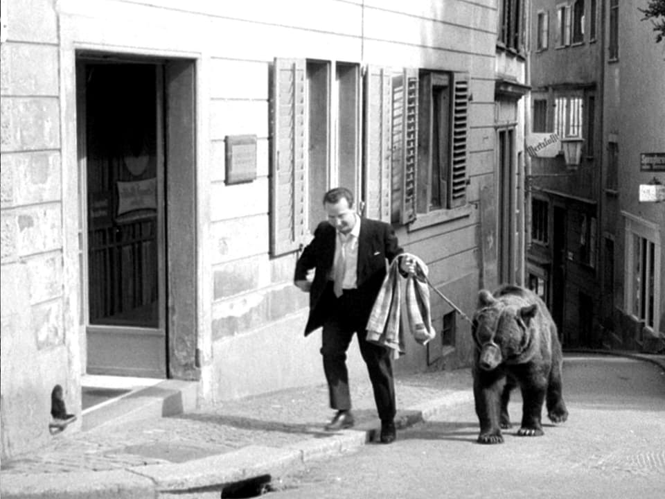 Ein Mann führt einen Braunbären an einer Leine in einer leeren Gasse spazieren.