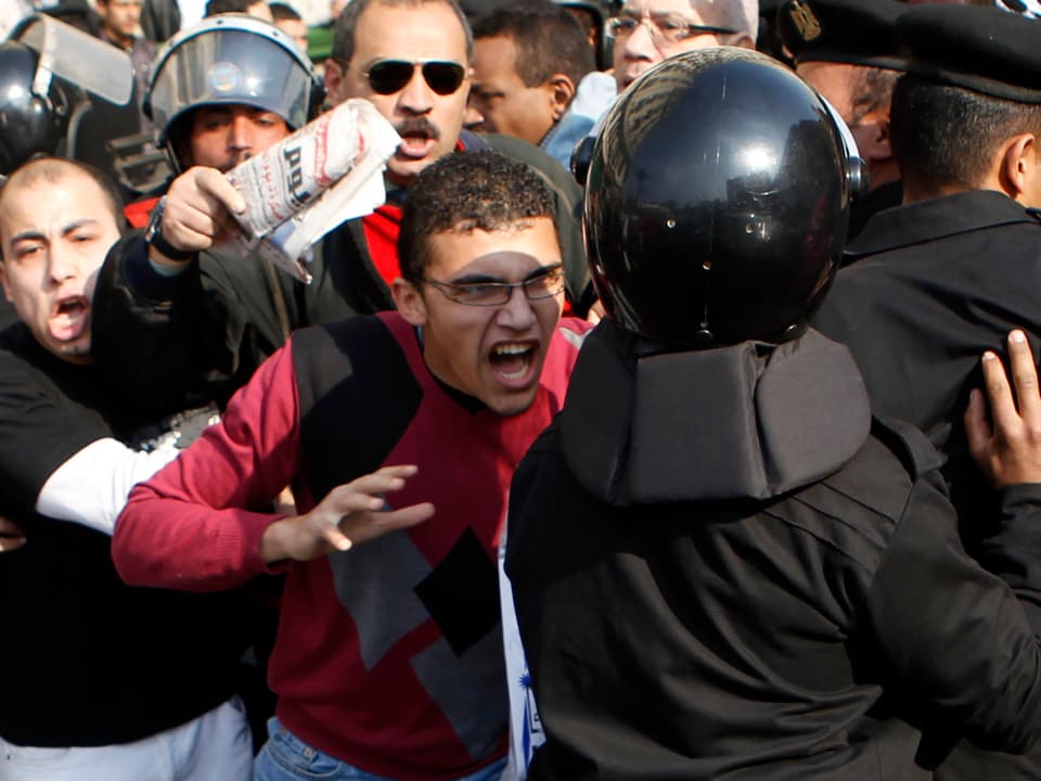 Ein Polizist in schwarzer Schutzkleidung und Helm schreitet gegen die wütende Masse in Kairo ein, ein junger Ägypter in bordaux-farbenem Pullover will sich auf den Polizisten stürzen. 